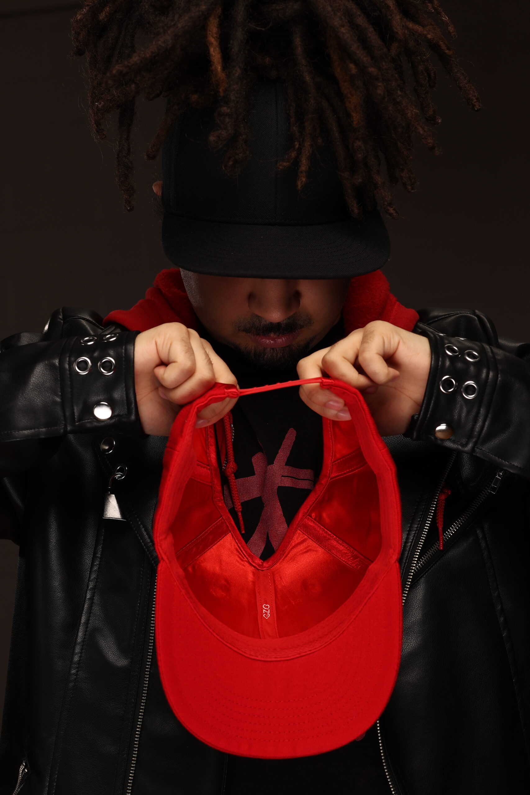 young man wearing a black leather jacket with his locs in a ponytail pulled through a black snap back baseball cap looks down at red, satin lined snap back made for locs, the back is removed in an A frame shape with the peak at the crown of the head, allowing protective hairstyles to be pulled up through the hat and off the neck.