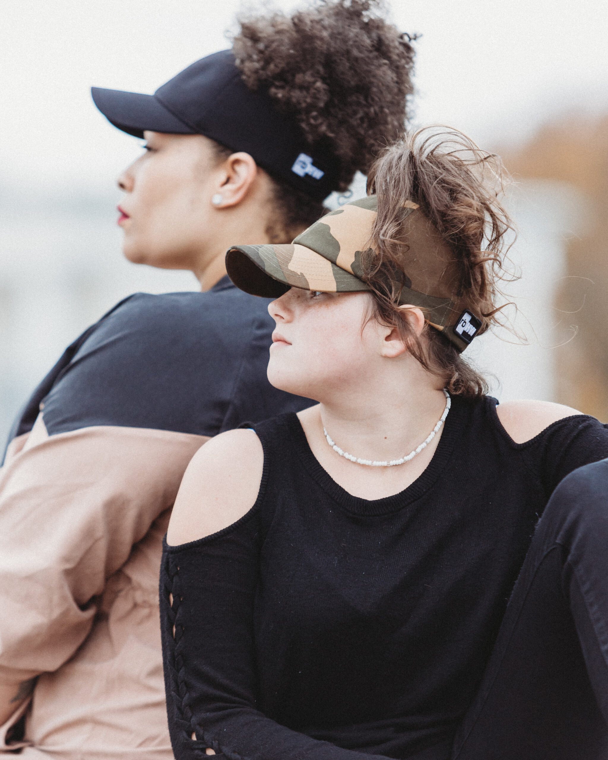 two women are shown on profile, looking into the distance, to the left. the woman in the background is a light-skinned black woman wearing a satin lined, black, super high ponytail baseball cap that allows her natural 4c hair to easily come through the top. the woman in the foreground is a younger, freckled white pre-teen wearing a satin lined, high ponytail, camouflage patterned baseball cap that allows her crown-height high ponytail of loose curls easily sit on the top of her head. both women's hats fit them neatly and without extra material.